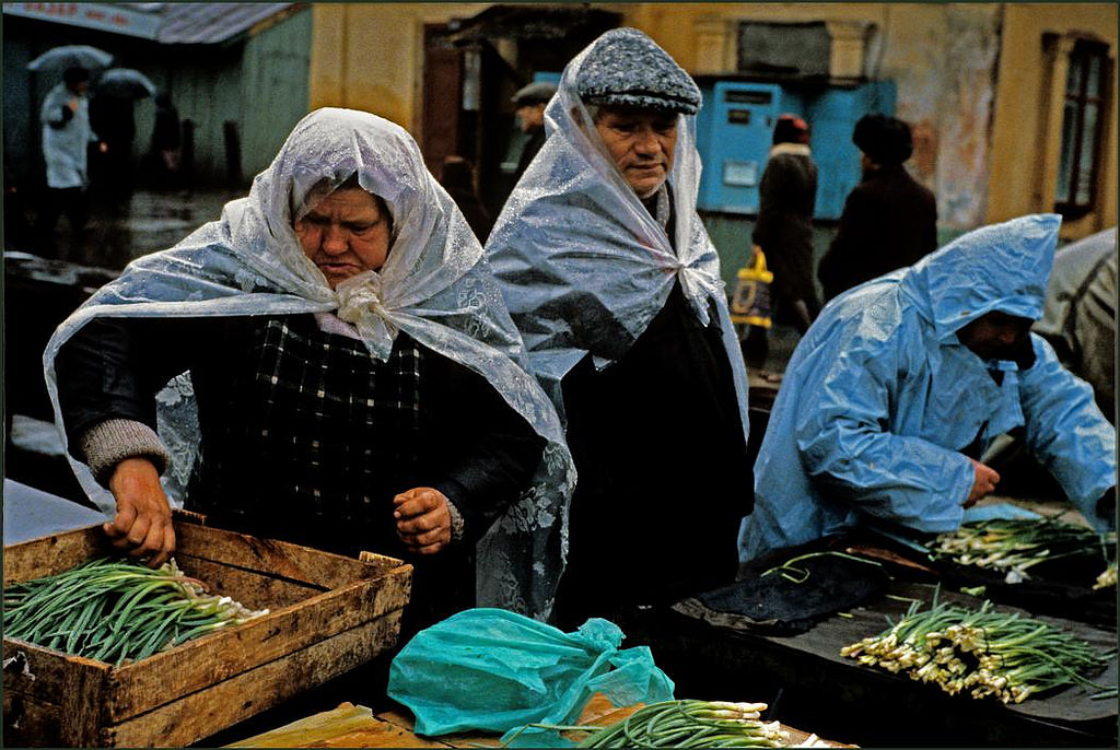 Men and women market stall holders shelter from heavy rain under plastic sheeting whilst sorting their onions.