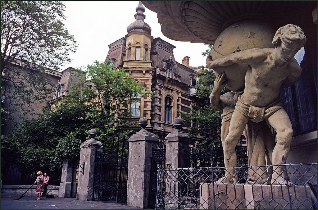 A woman sweeps the street in front of a grand old house with a statue of two loin cloth wearing men holding up the world.