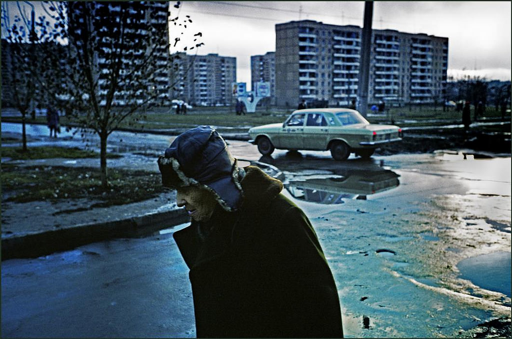 An elderly man dressed against the wet weather makes his way through the puddles in a dreary suburb of tower block flats whilst a Russian made car passes in the background.