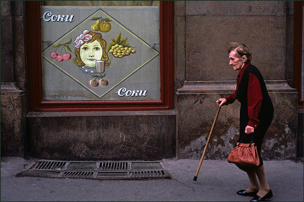 Elderly lady with walking stick passing a window advertising a fruit drink.