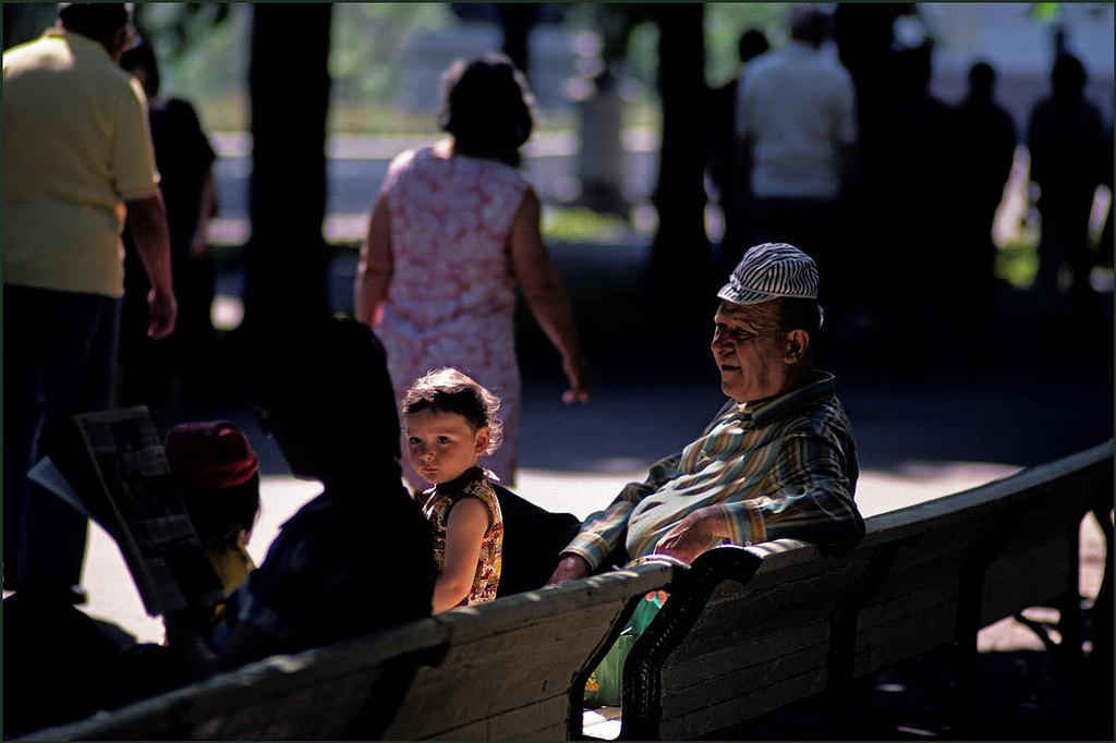 Family sitting on a bench in the park catch the last rays of the sun whilst the man wears his grandaughters hat and the mother reads.