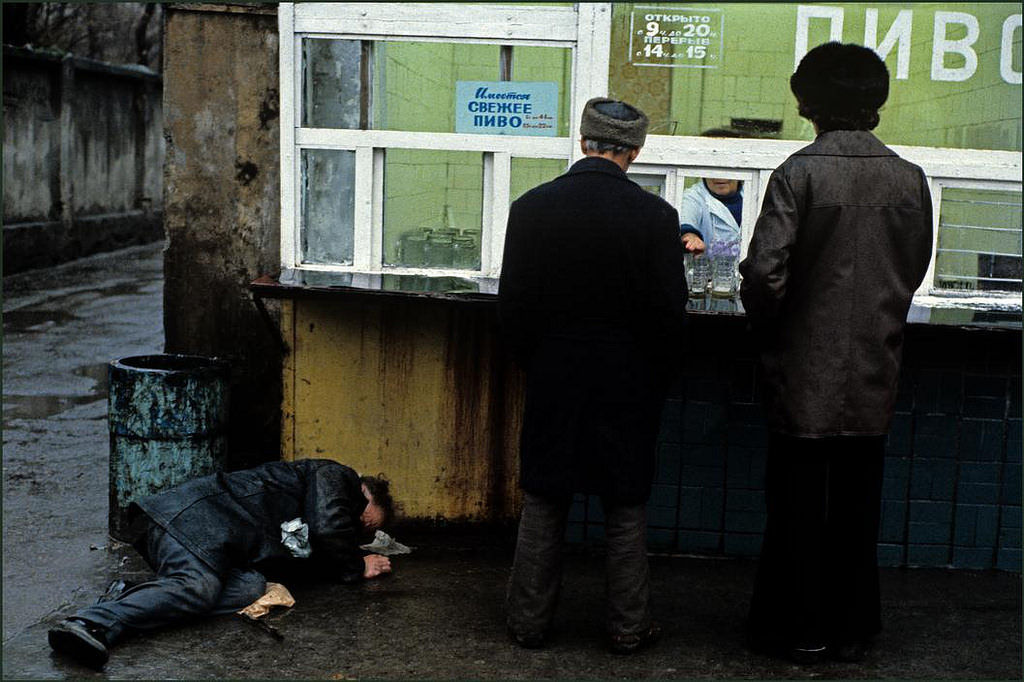 Two men buying beer from a street kiosk in the rain whilst another lays drunk on the ground.