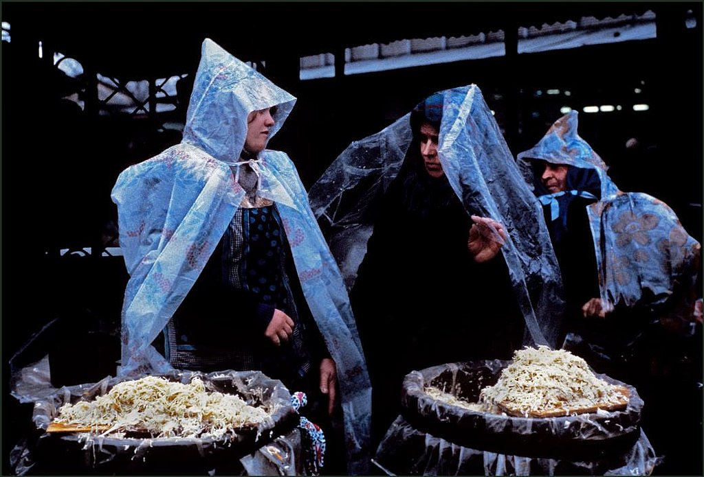 Women tending a market stall shelter from heavy rain under plastic raincoats.