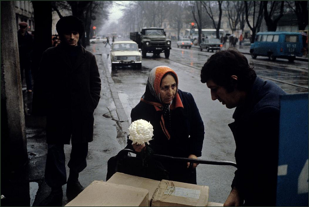 A woman trying to sell a flower to a man unloading packages in the rain on a shopping street.