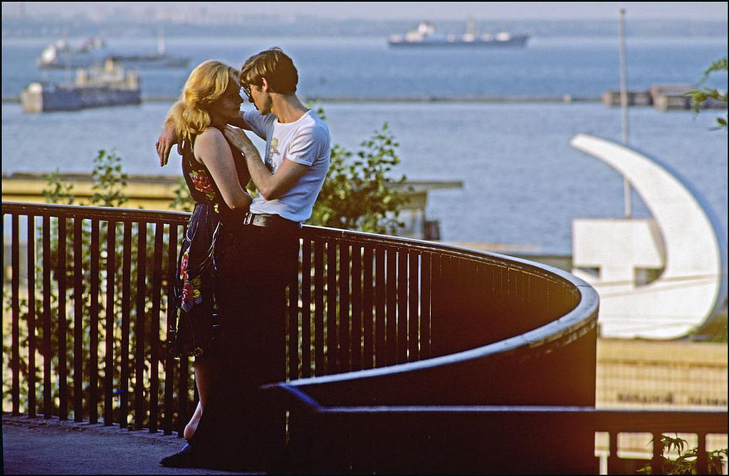 A couple of lovers meet on a balcony overlooking the port with a hammer and sickle sign and ships in the background.
