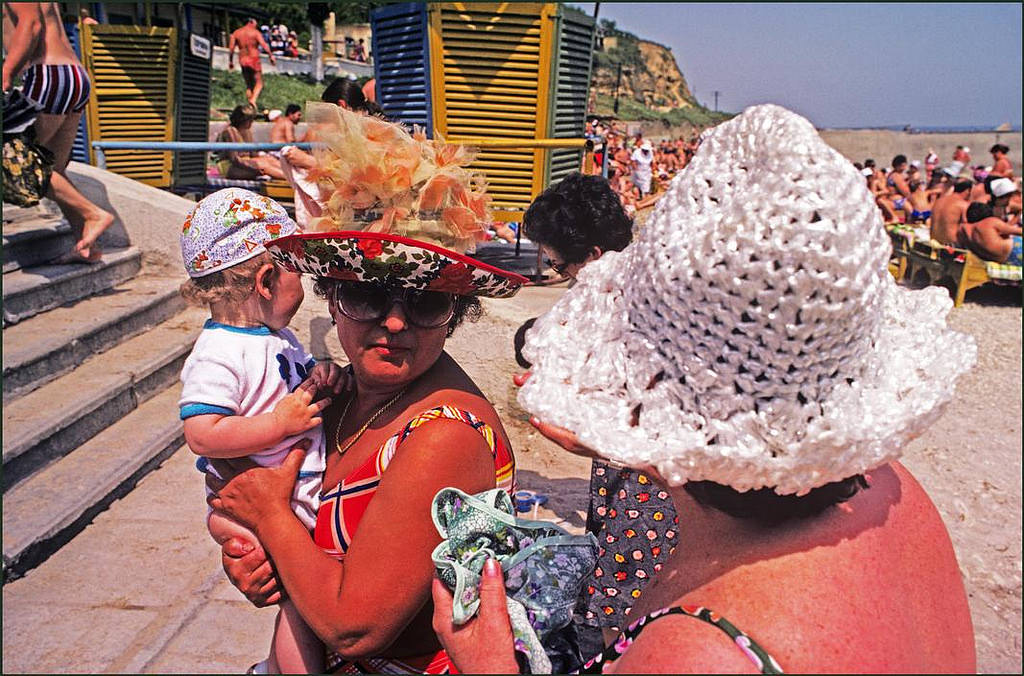 A woman in an amazingly decorative sun hat holds her child whilst chatting to another lady on the beach.