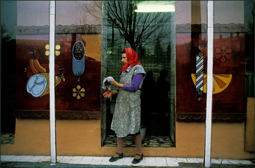 Woman cleaning the window of a grocery store with fruit and vegetable paintings.