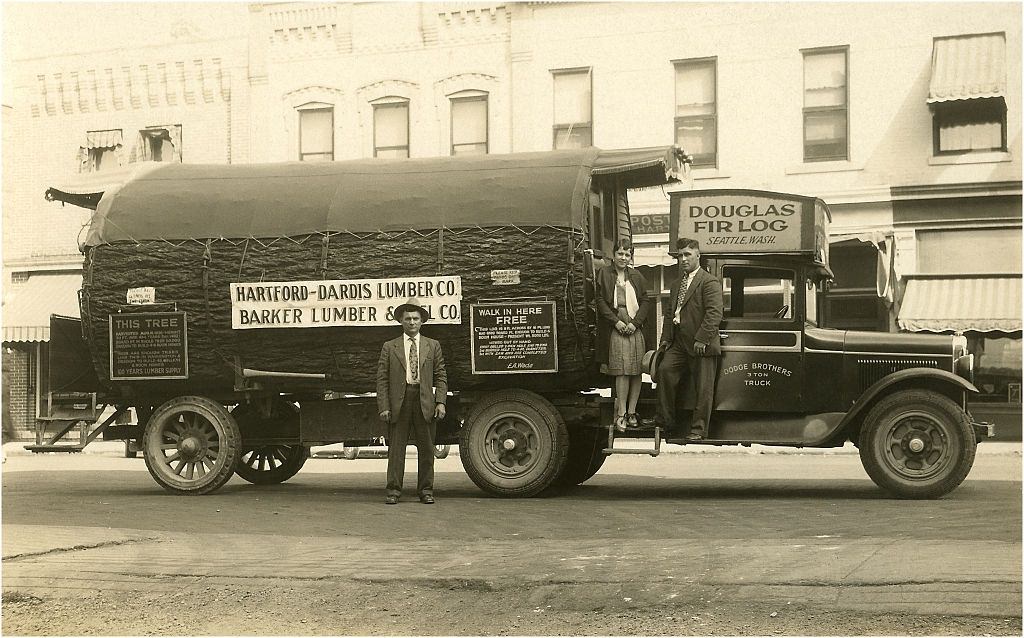 Three people standing with a giant Douglas fir log converted to a structure and mounted on a truck, ‘Barker Lumber & Fuel Co., Seattle, 1930s.