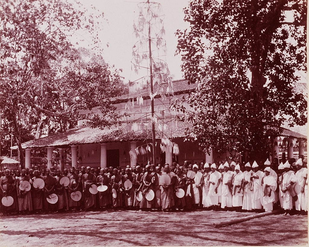 A group of men dressed in white robes standing next to another group of men, several of whom carry straw fans, Sri Lanka, 1880s.
