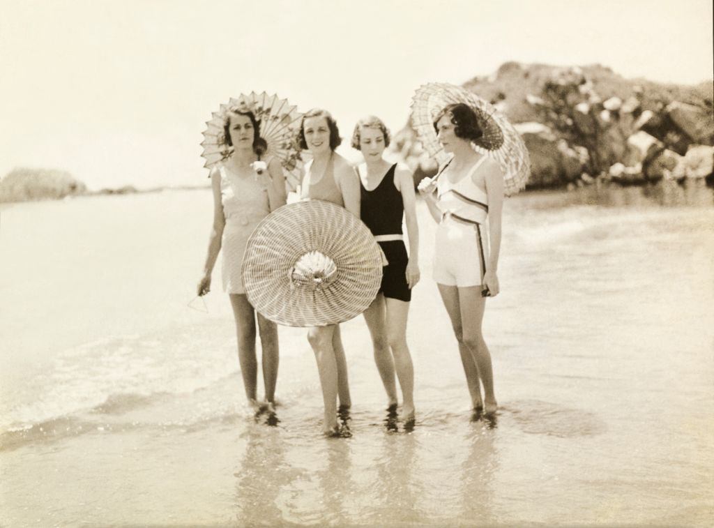 Mrs. Douglas, Mrs. Cushing, Mrs. Phipps, and Mrs. Stevens holding parasols and standing with their feet in water at the beach, in Newport, Rhode Island.