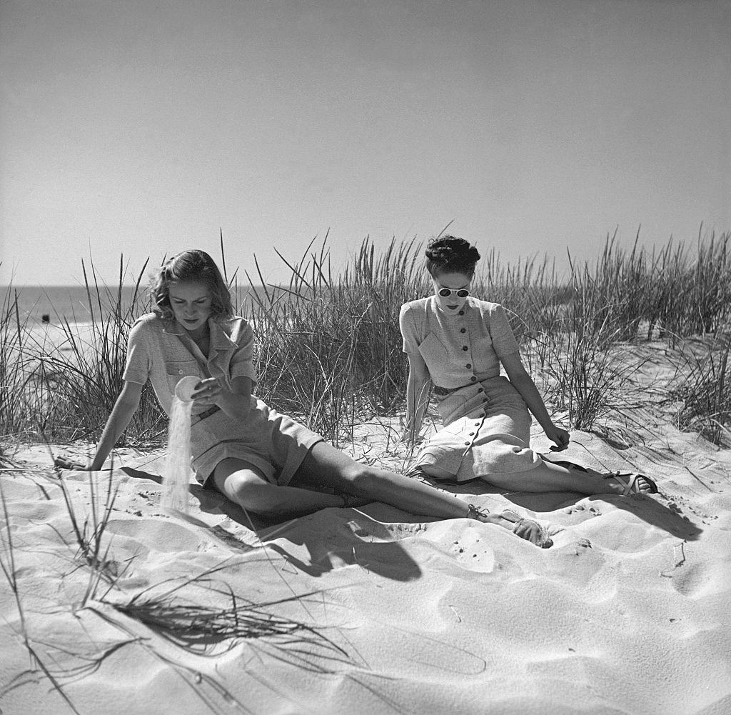 Two models sitting on grassy sand dunes with the ocean in distance, 1937.