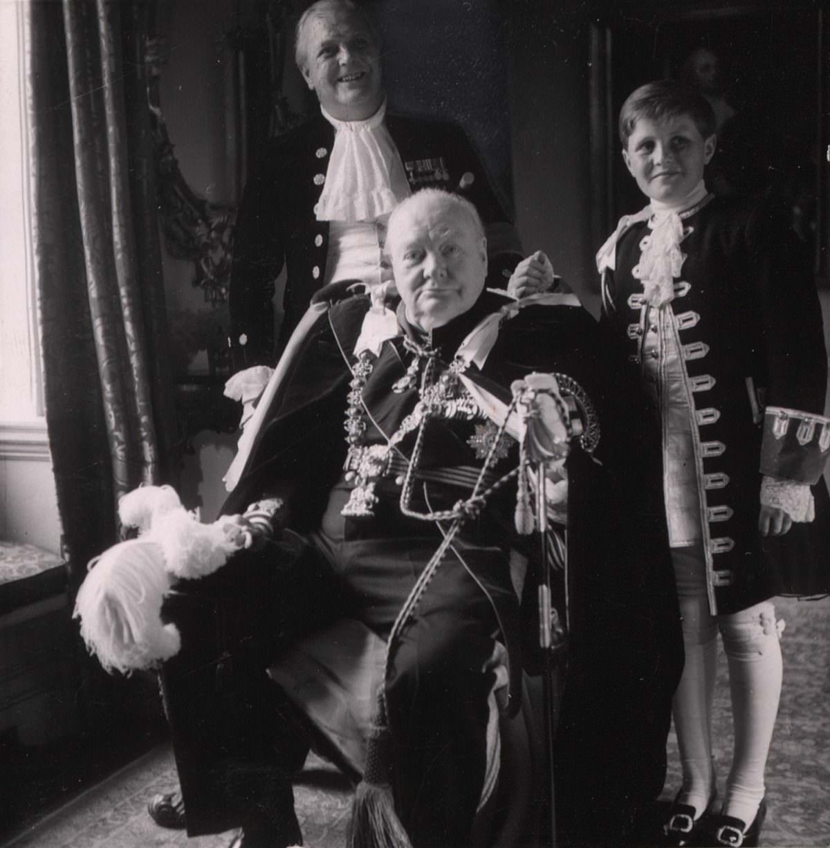 Winston Churchill, full-length portrait, seated, facing front, with his son and grandson standing behind him, wearing coronation robes.