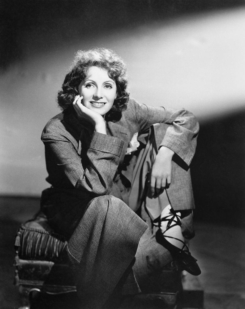 Greta Garbo seated on an ottoman, wearing a large business suit and sporting ballet shoes, 1930s.