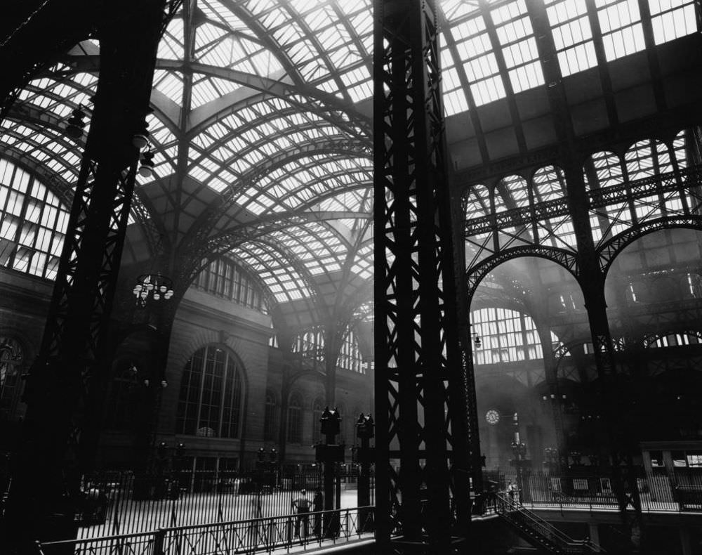 Penn Station, Interior, Manhattan. Massive steel uprights in the center and to the left, with lighter steel tracery and windows above; men stand near stairway to trains.