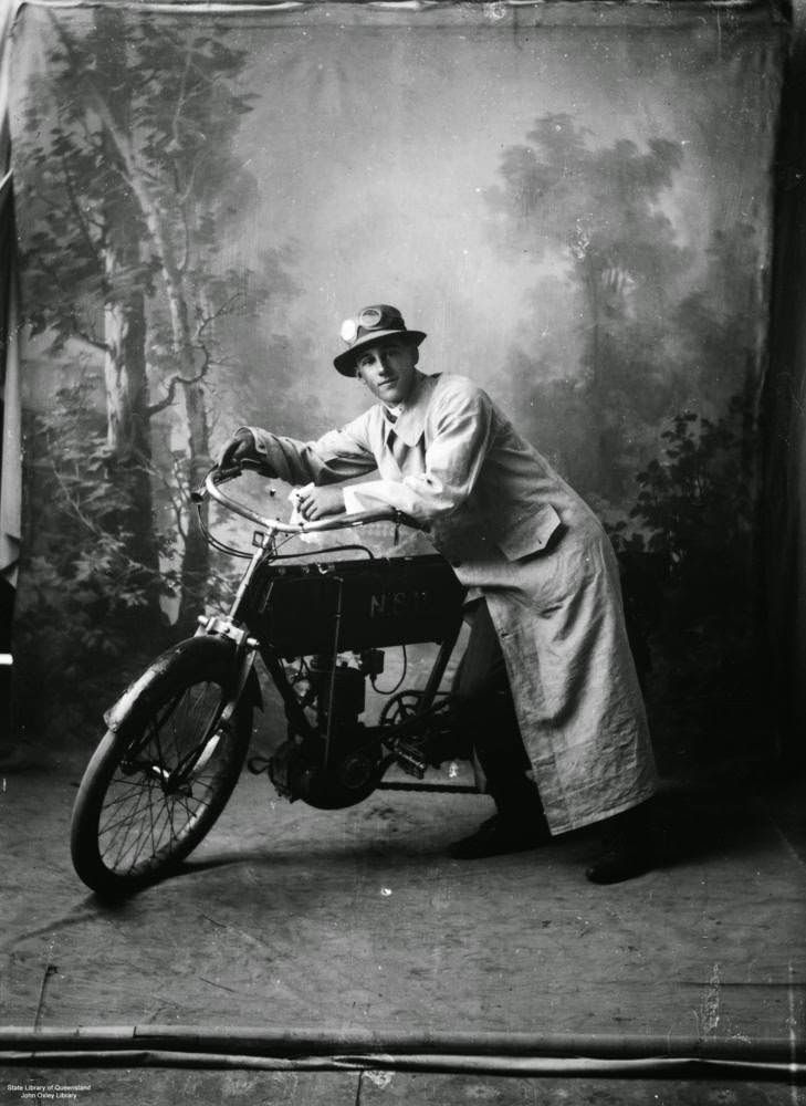 Man dressed in a long dustcoat, hat and goggles with a motorcycle standing in front of a painted backdrop in a studio, 1935.