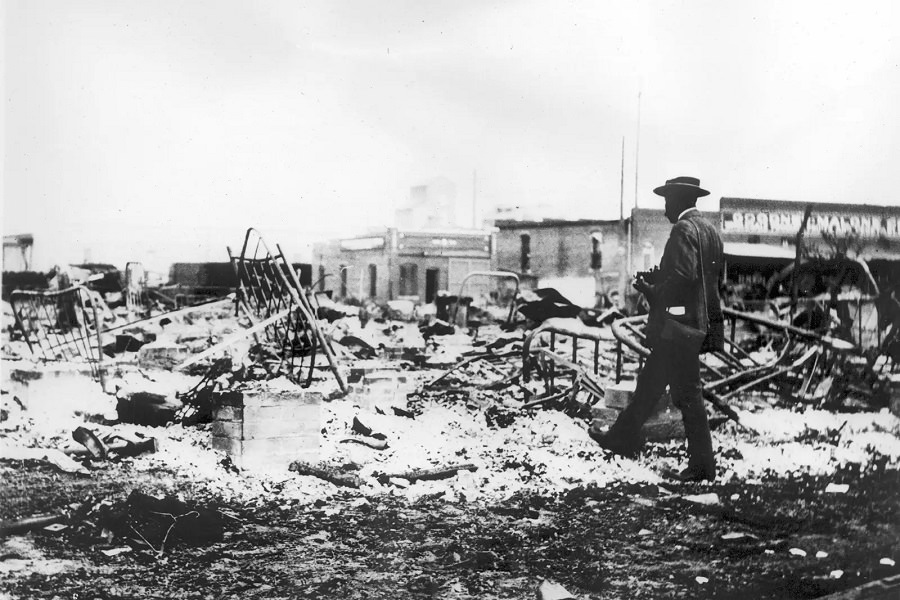A Black man with a camera inspects the skeletons of iron beds rising above the ashes of a burned-out block in Tulsa, June 1921.