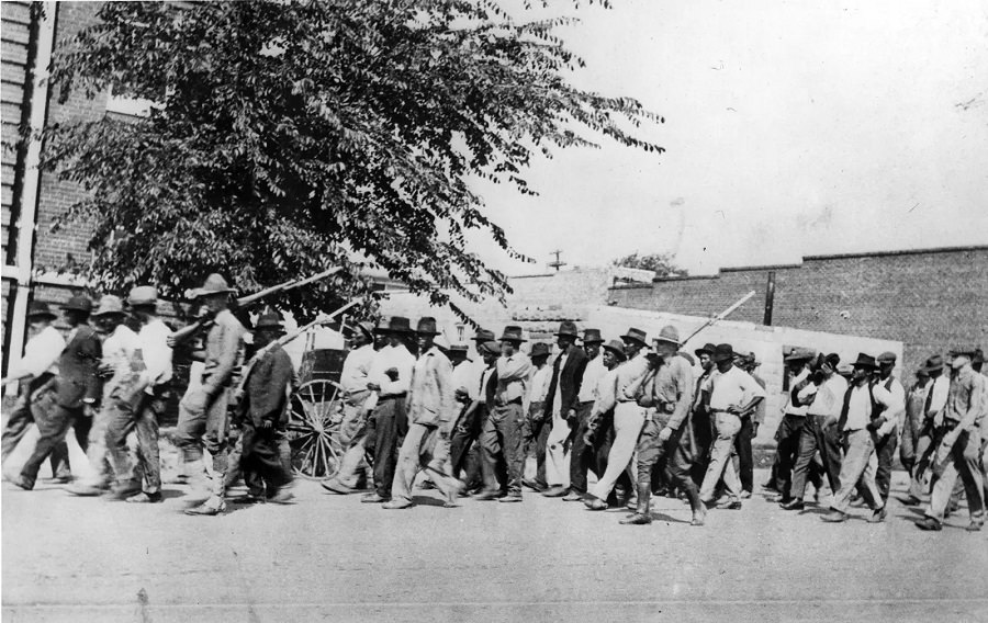 A group of National Guard troops, carrying rifles with bayonets attached, escort unarmed Black men to the detention center at the Tulsa convention hall in June 1921.