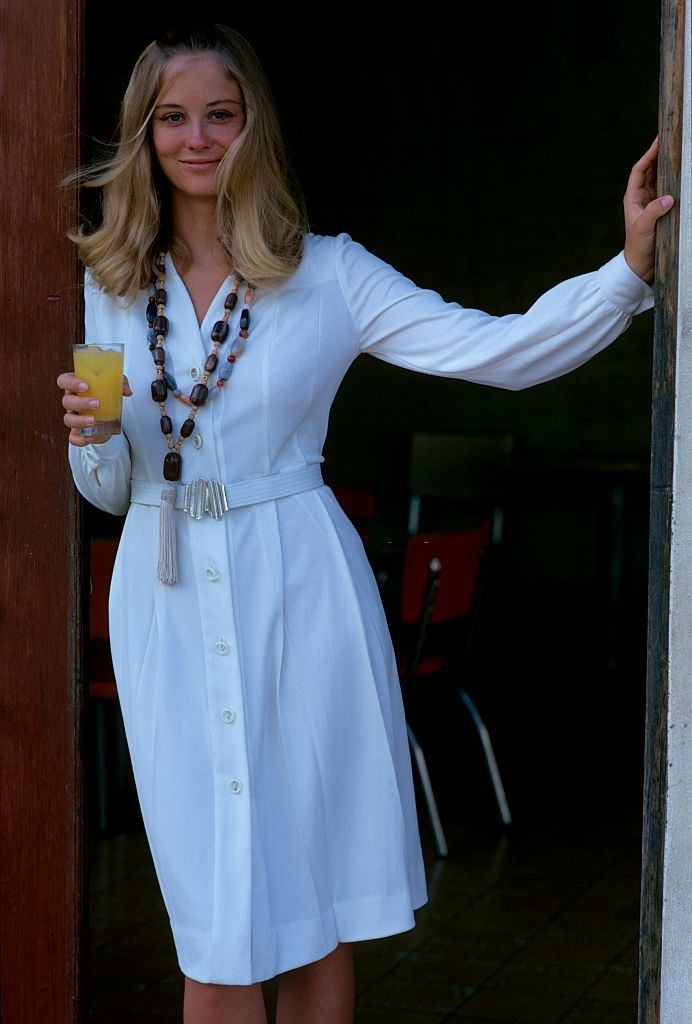 Cybill Shepherd holding a glass of orange juice, wearing a white dress with a pleated skirt, Glamour 1970.