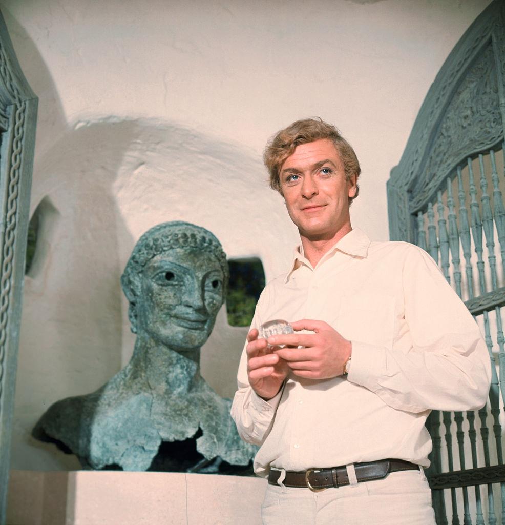 Michael Caine stands next to a bronze Etruscan bust, 1960s.