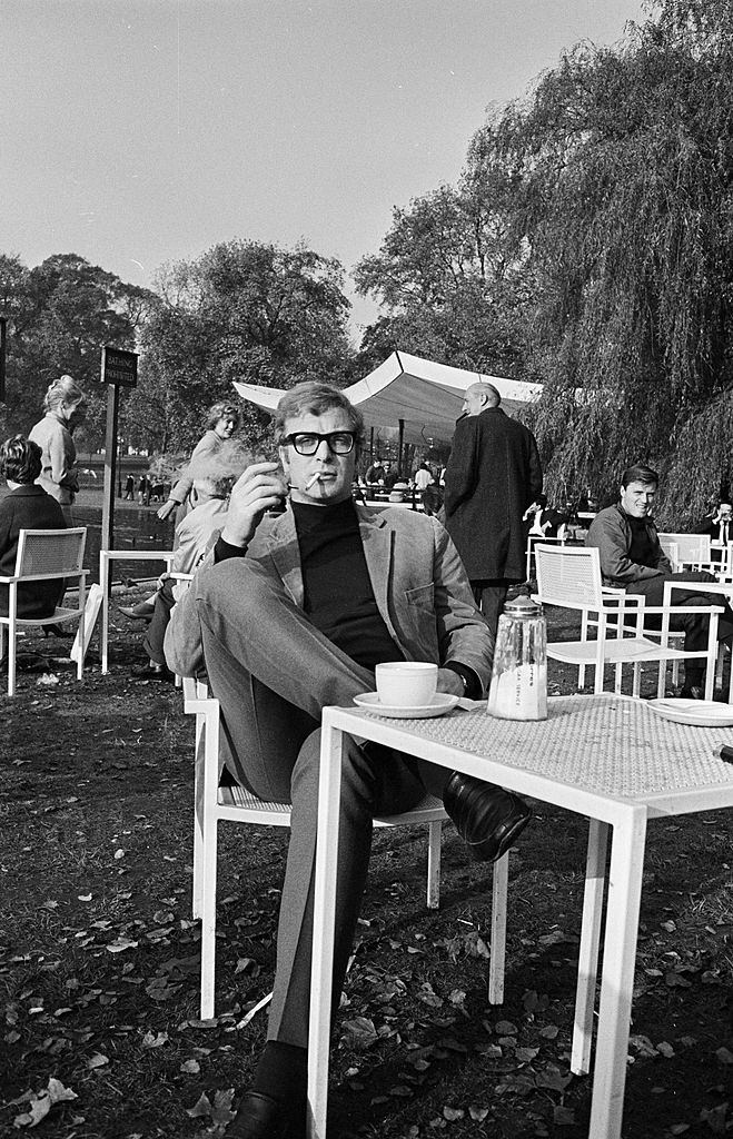 Michael Caine with a cigarette in his mouth, sitting cross-legged on a terrace in a park, 1965.