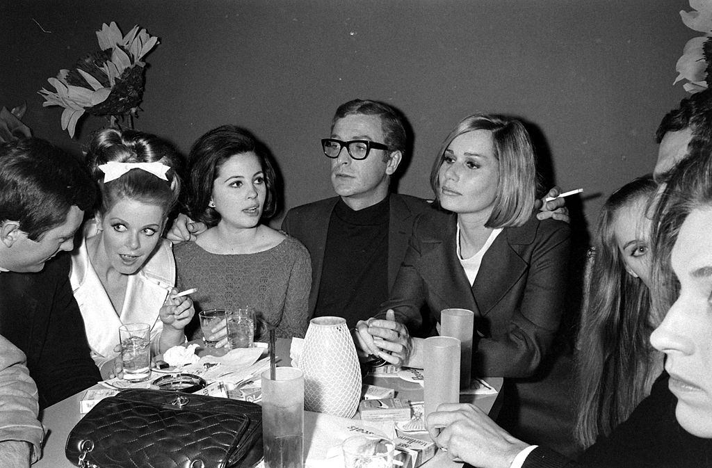 Michael Caine smoking a cigarette and dining with friends, including the actress Sally Kellerman, 1966.