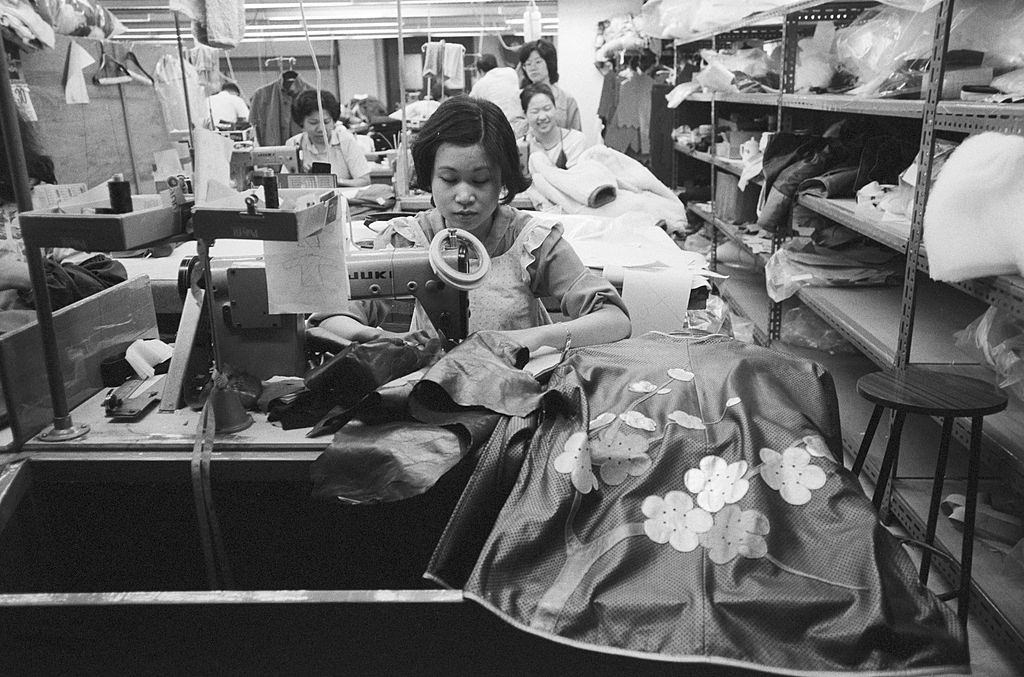 A seamstress works in front of her sewing machine arranged perpendicular to a shelf. Hong Kong, 1981.