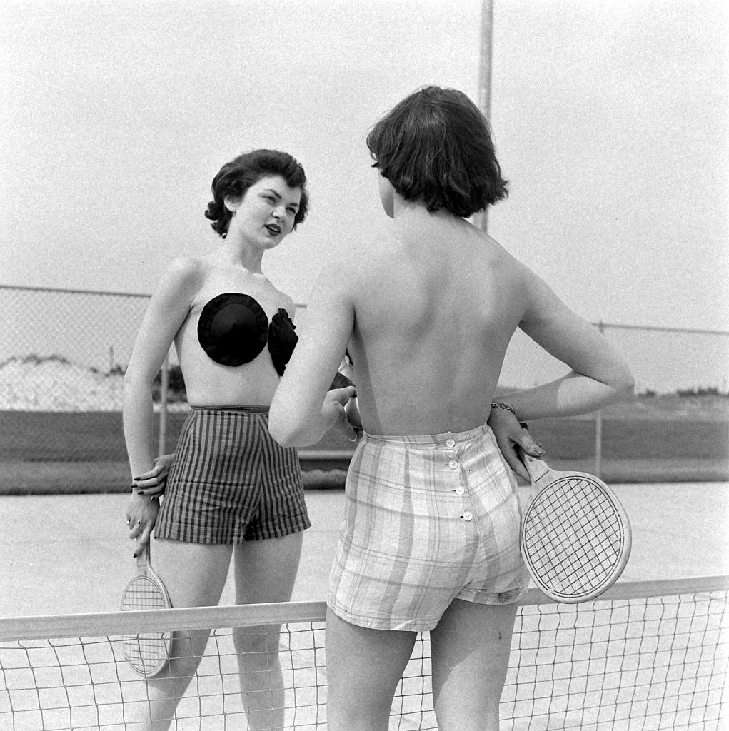 Two models, dressed in shorts and 'Poses,' an adhesive, strapless brassiere designed by Charles L Langs, hold fake tennis rackets as they talk over a net, Jones Beach, New York, May 1949.