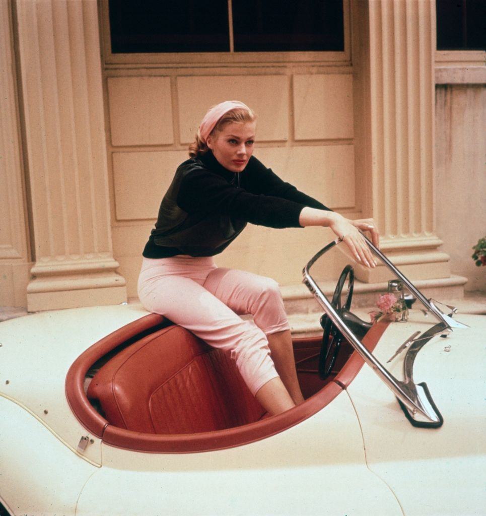 Anita Ekberg sitting on the trunk of a cream convertible with her feet in its red leather interior, 1955.