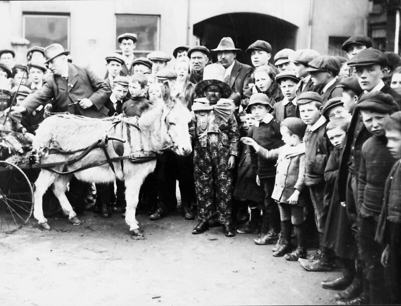A crowd of people round a clown and a donkey, 1910.