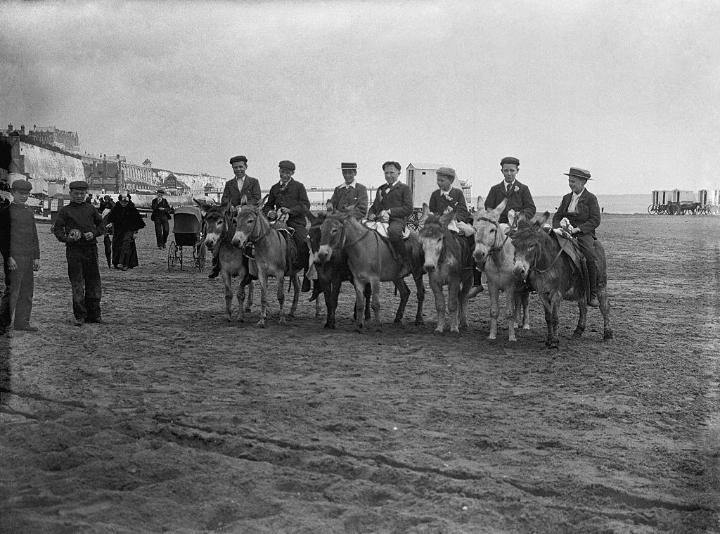 Young group of boys in cloth caps and boaters sitting on donkeys on a sandy beach Ramsgate Kent.