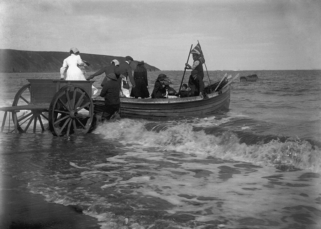Smartly dressed family group being helped from a two wheeled cart into a coastal rowing boat on an English beach circa 1900.