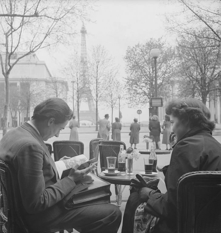 An unidentified couple sit at a sidwalk cafe near the Eiffel Tower and read. Each has a bottle of Coca-Cola in front of them.