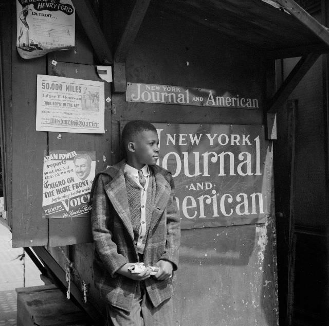 A newsboy standing by his newsstand holding a magazine, 1943.