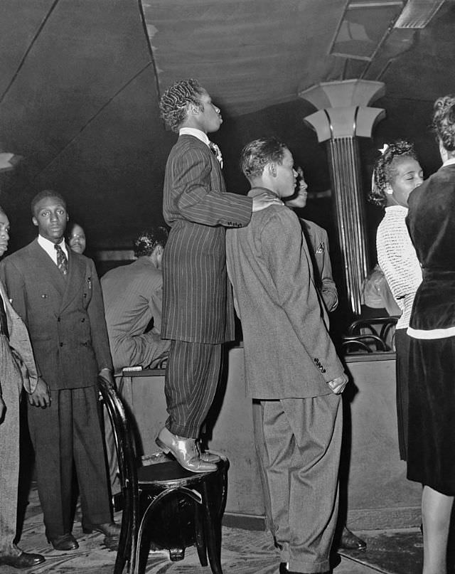 A short man, wearing a zoot suit, stands on a chair to get a better view of the dance floor at the Savoy Ballroom, 1940s.