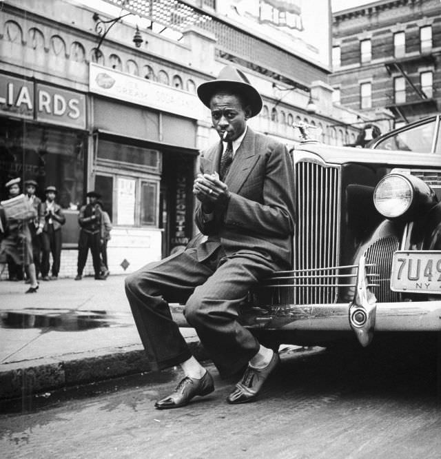 Baseball player Satchel Paige, lighting his cigarette while sitting on the front bumper of a large car, 1941.