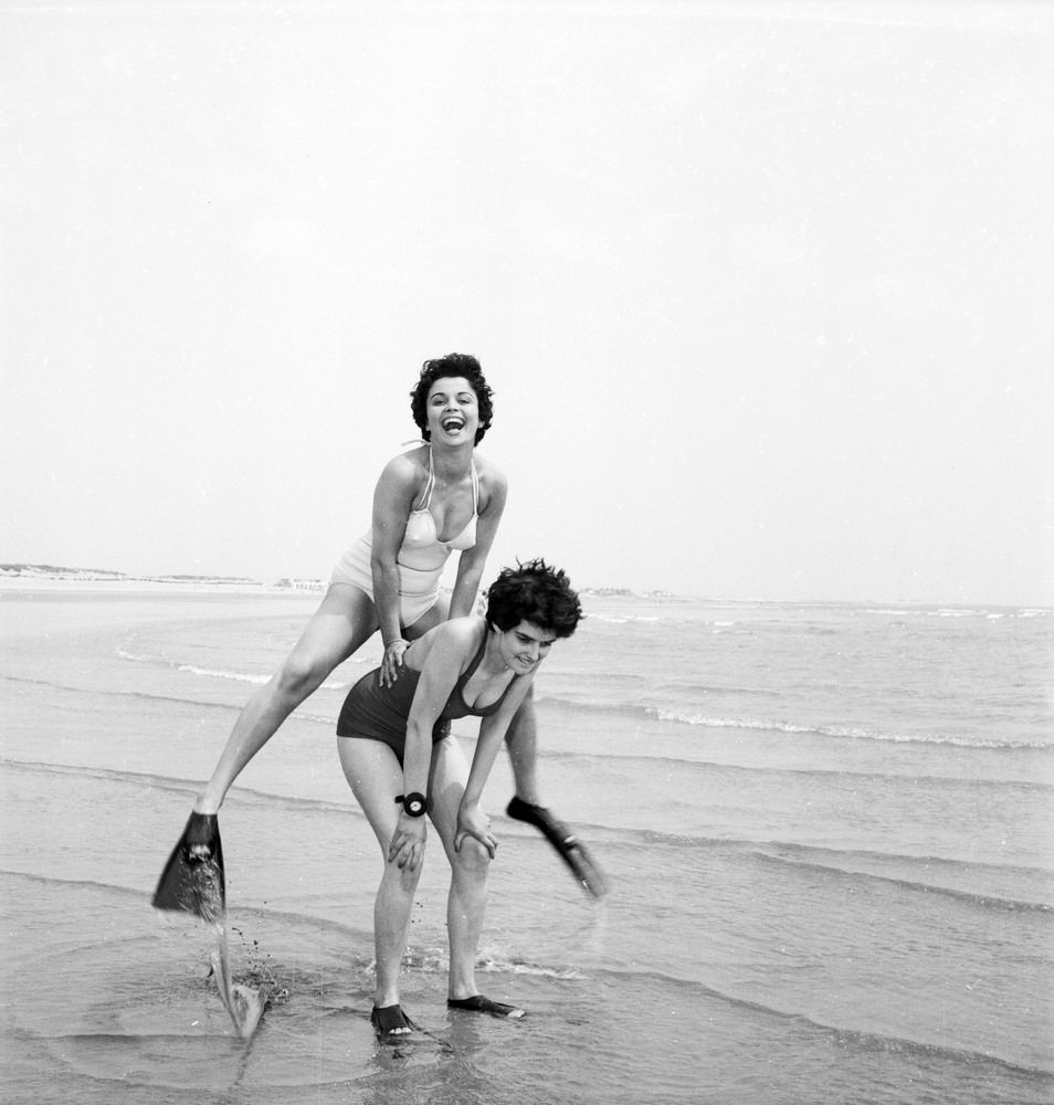 Two women wearing fins play leap-frog on the beach, 1955.