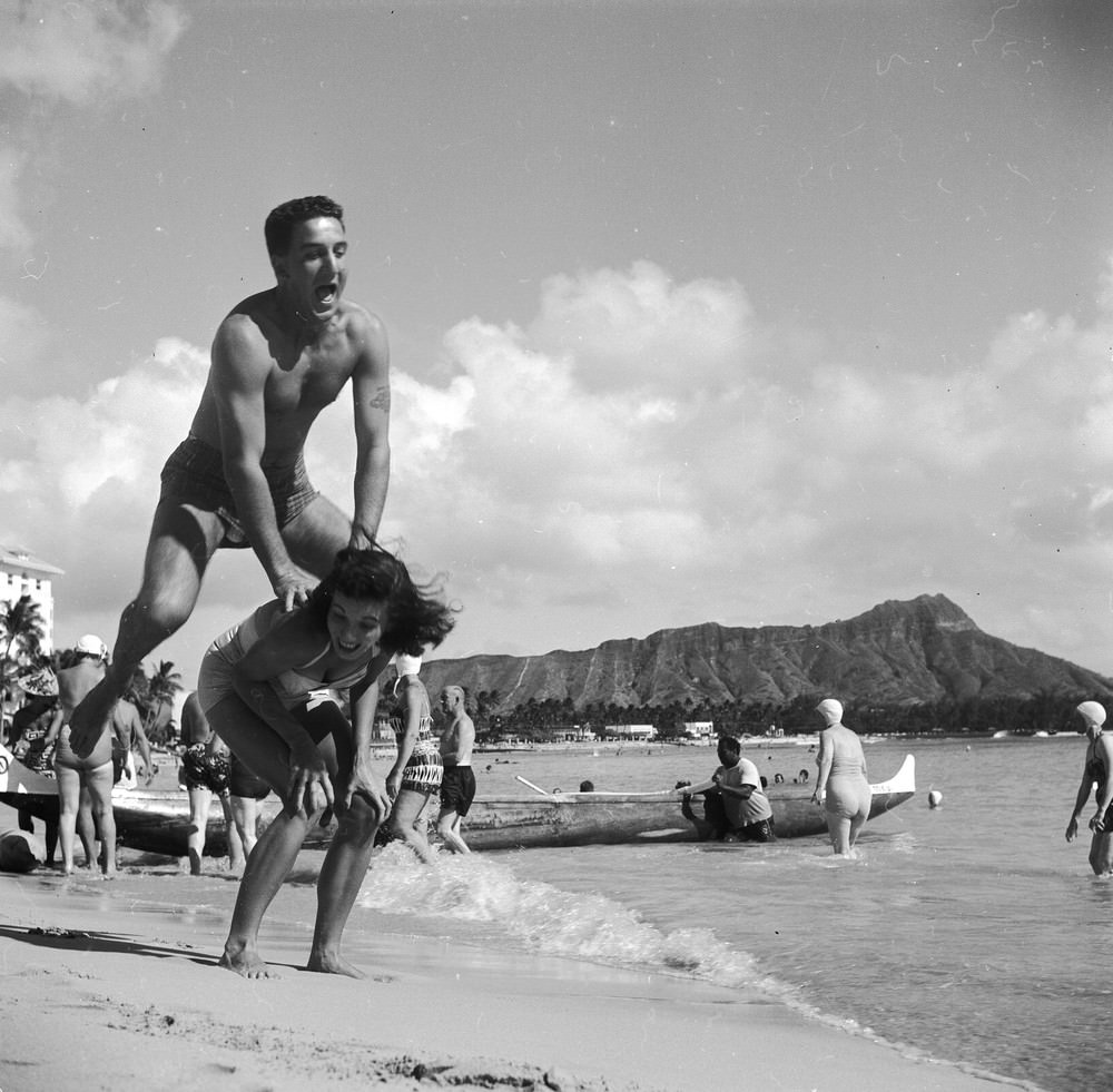 Bert and Ellie Lang, a young American couple on their honeymoon in Hawaii play leap-frog on Honolulu's Waikiki beach. In the background is the extinct volcanic crater of Diamond Head, circa 1955.