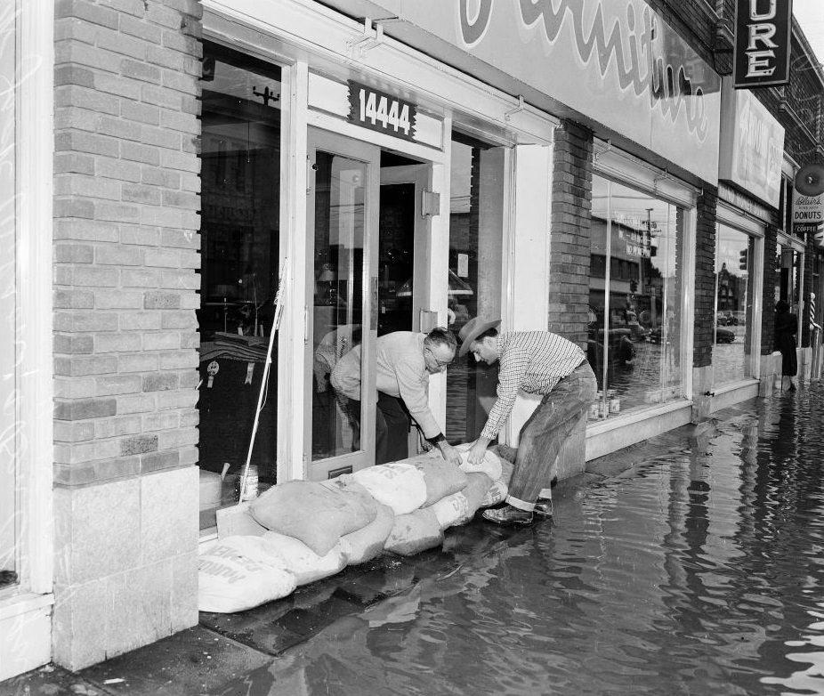 Man helping a shopkeeper by putting a sand bag in front of the store, 1952.