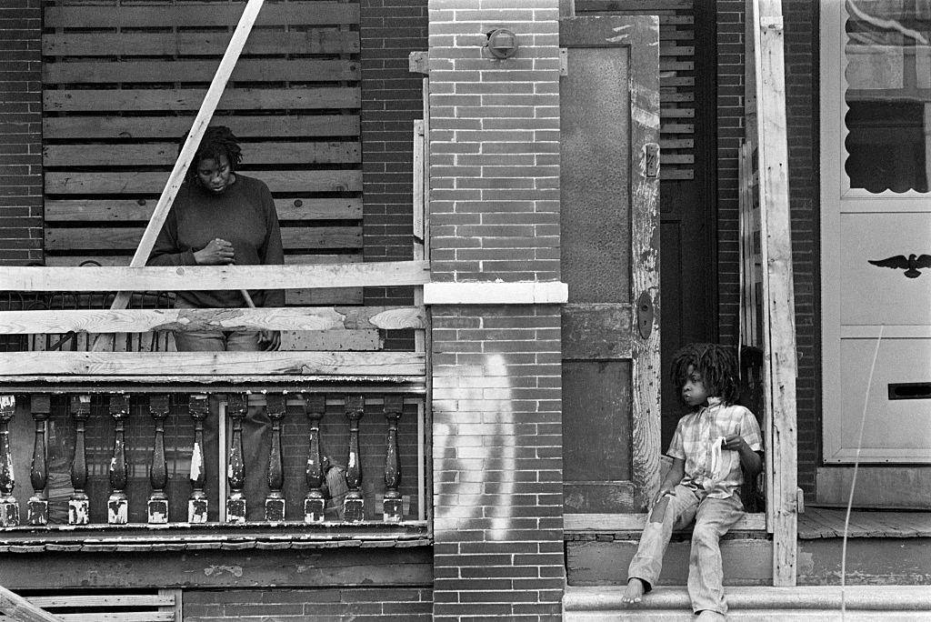 A woman cleans her porch while a child in dreadlocks sits on the steps eating lunch.