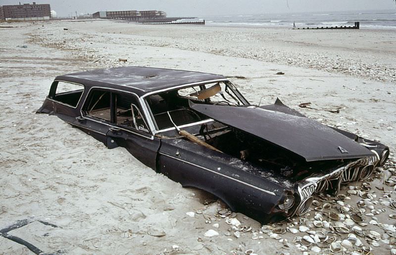 Half buried early 1960s Dodge Polara station wagon on the beach at the ocean side of Breezy Point in Queens, an abandoned apartment building at left distance, 1973