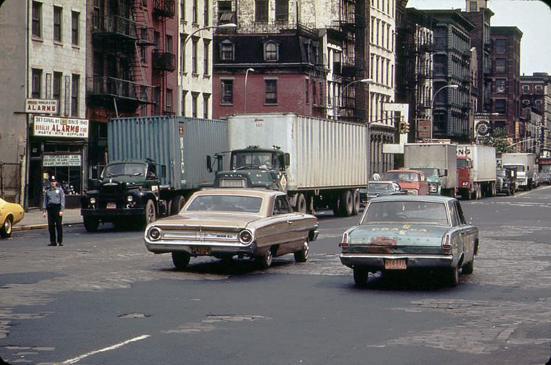 A 1960s Ford Galaxie and a battered early 1960s Plymouth Valiant cruise along Canal Street with peeling pavement, 1973