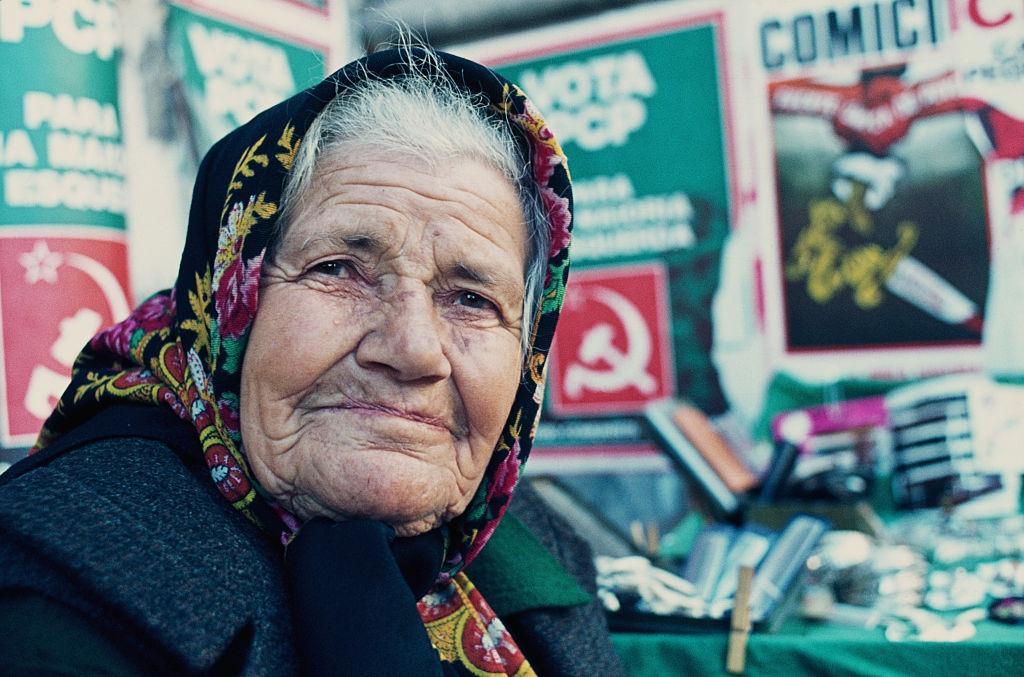 An elderly woman in front of a wall covered in Portuguese Communist Party (PCP) billboard posters, Portugal, circa 1970.