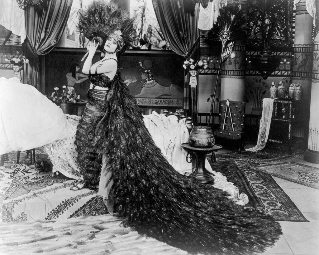 Theda Bara holds a peacock feather fan next to a bed, 1917.