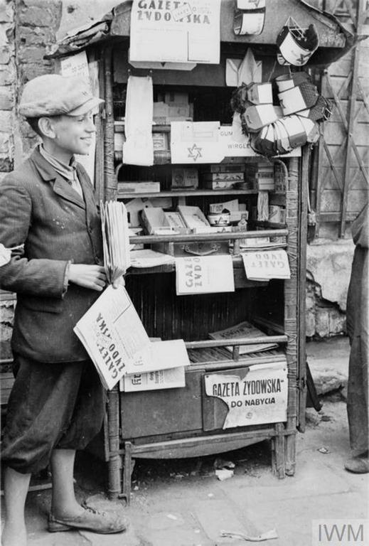 A young and cheerful seller of newspapers and armbands running his stall in the street of the ghetto (possibly Muranowski Square). The title of the newspaper for sale is “Gazeta Żydowska – Jewish Gazette”. He also advertises Wrigley’s chewing gum for sale, mispelled as Wirgley’s.