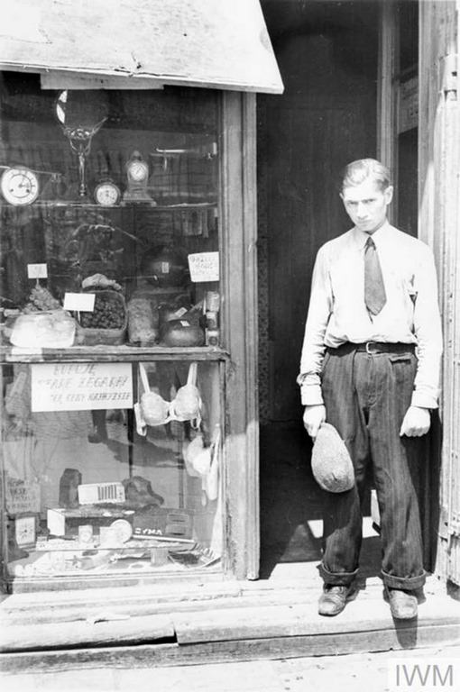 A young man in the doorway of a shop in the ghetto. Note he has taken his hat off to comply with the German order to remove headwear in the presence of German personnel. The shop offers fresh eggs, sweets and watches. The sign in the window reads – “I buy old watches for top prices”.