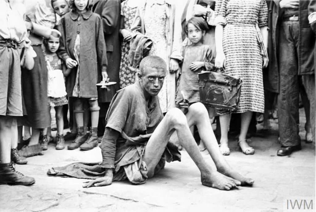 An emaciated boy sitting on a pavement. Note a crowd of pedestrians around him, including children with toys.