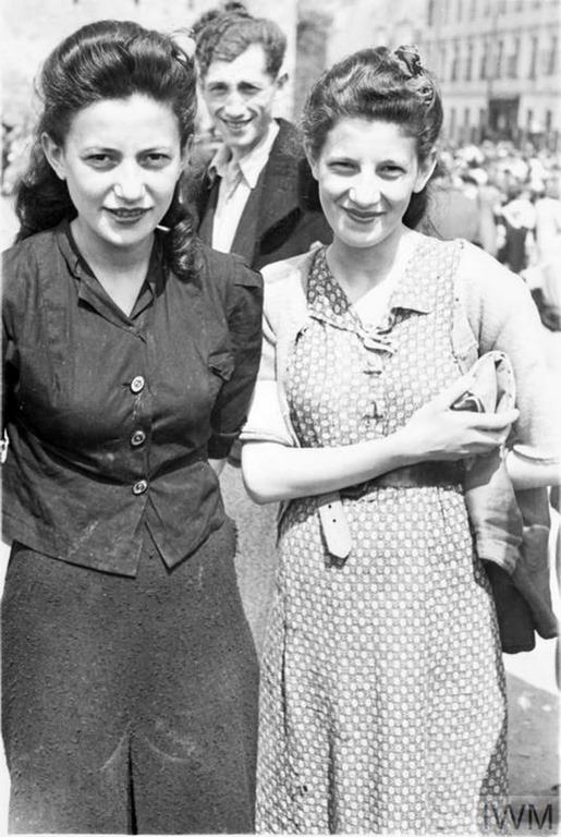 Two well dressed women, most likely sisters, posing for a photograph in a street market.