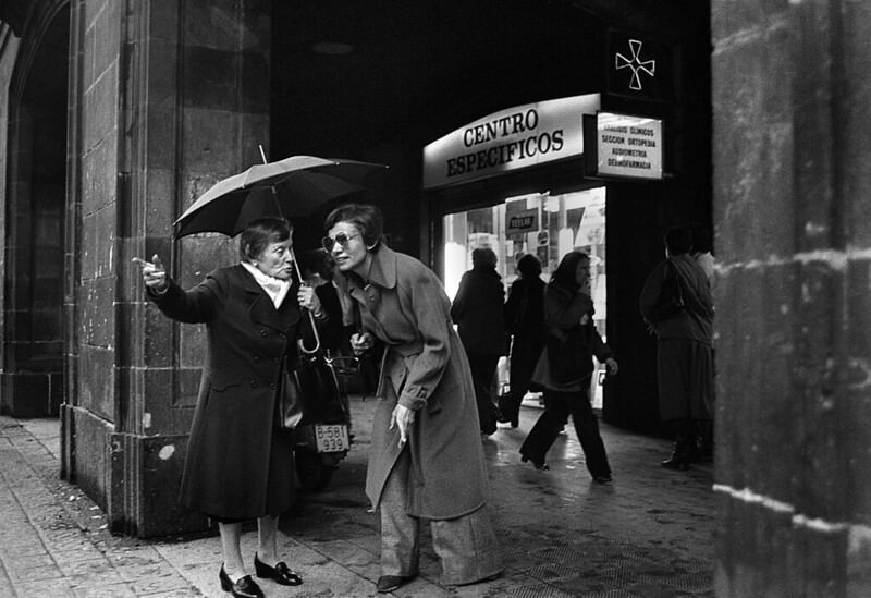 A woman in the neighborhood, with an umbrella, points in the direction of a street at a foreign woman. Barcelona, 1979.