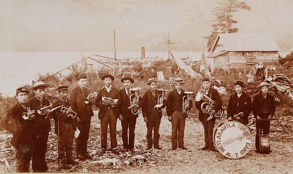 Members of the Metlakahtla Cornet band, a group of Native American brass musicians, pose for a picture next to buildings by the shore in an Alaska town, ca. 1892.