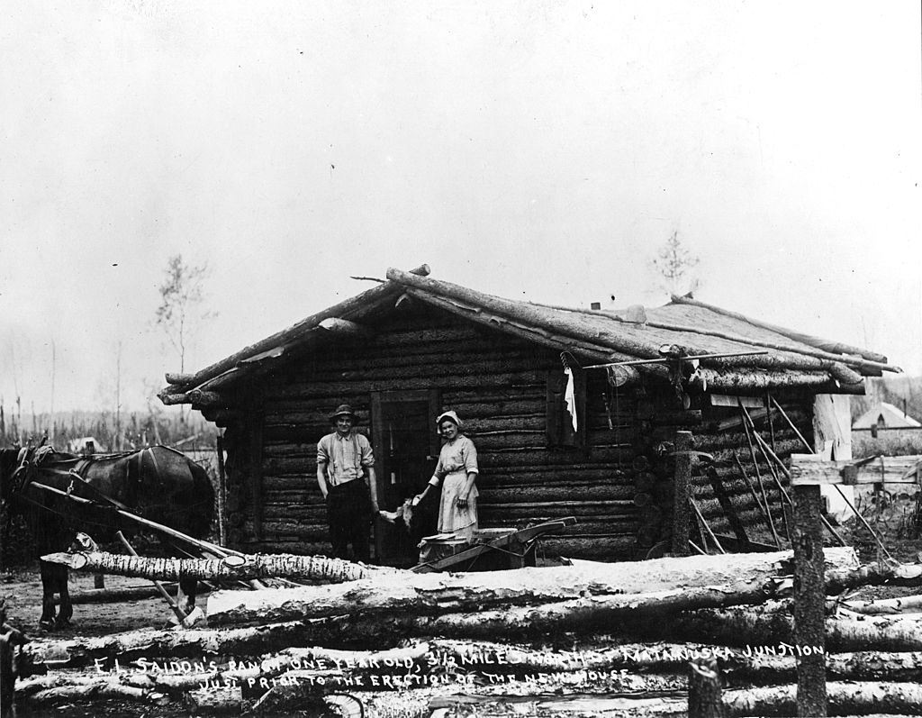 Man and woman as they pose at the doorway of a log cabin, Alaska, 1860s